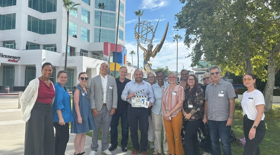 The Councilmember, holding a slate (clapper) poses with officers of teh Television Academy outside their headquarters, in front of a statue of the Emmy Award.